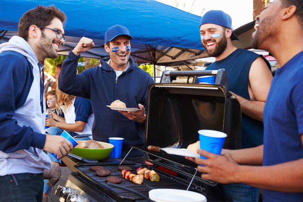 group of men gathered around a grill at a football tailgate