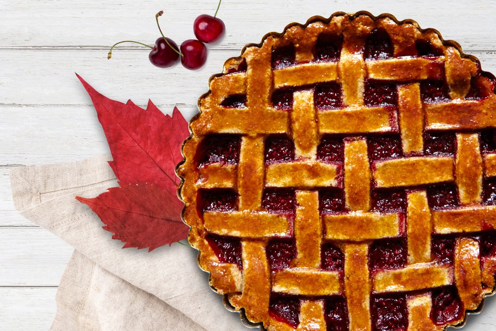 cherry pie on distressed, white wood table with red leaves, napkin, and cherries