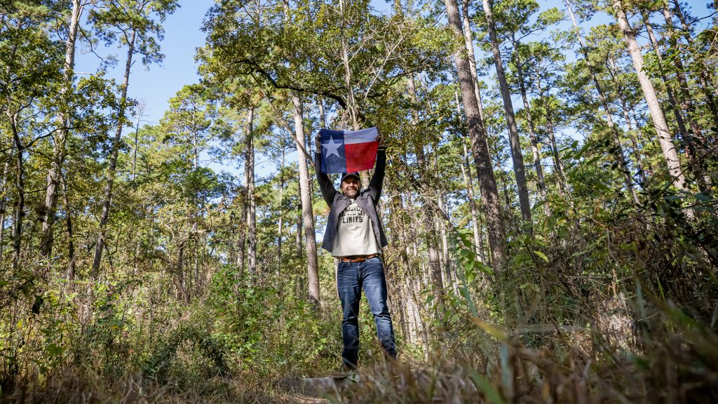 Chet Garner holding Texas flag in wooded area.