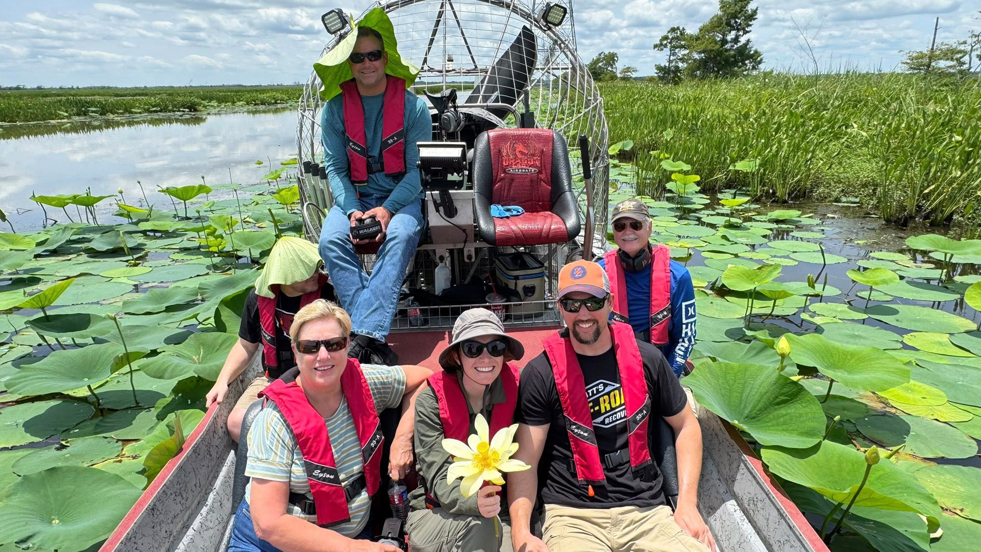 Group of people sitting in an air boat on the water surrounded by big lily pads.