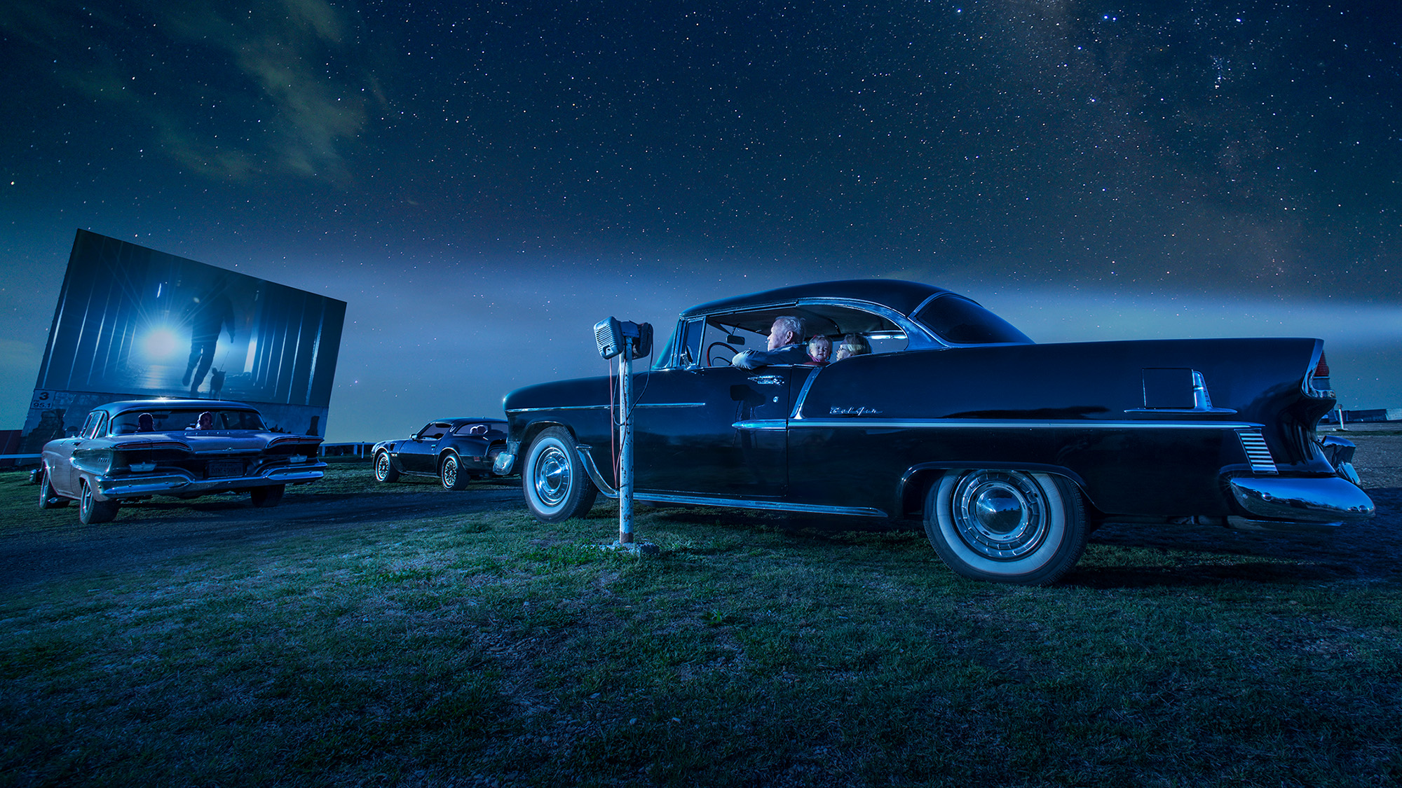 People in a car watching a movie at a drive-in theater at night.