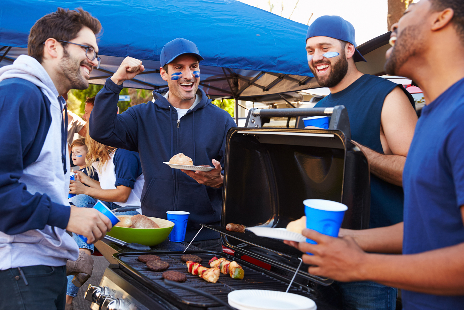 group of men gathered around a grill at a football tailgate