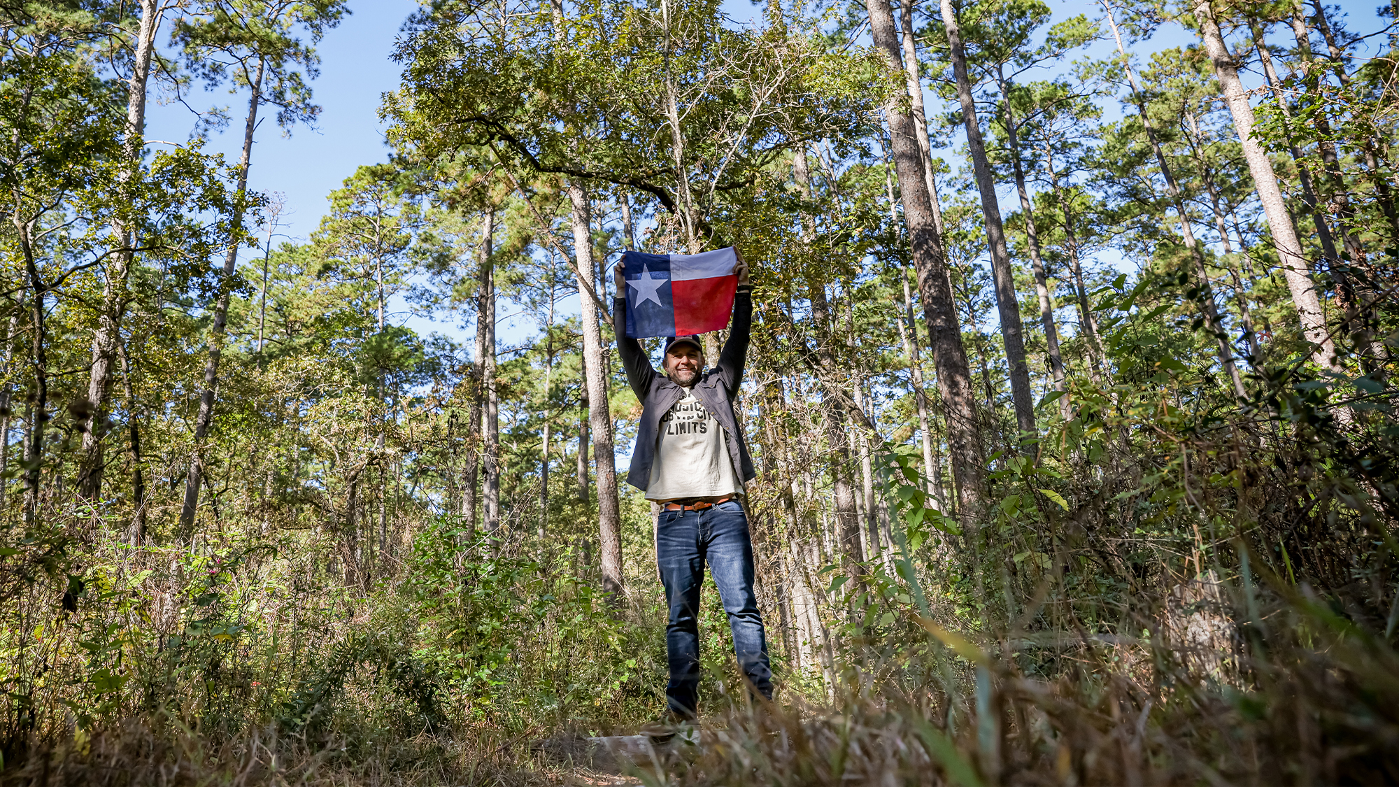 Chet Garner holding Texas flag in wooded area.